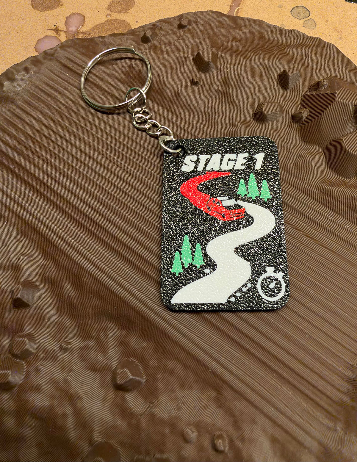 Svart nyckelring STAGE 1 med slingrande grusväg, röd rallybil, granar och tidtagarur